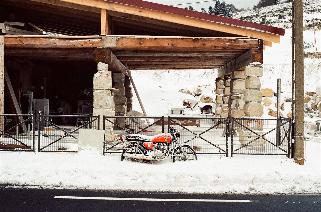 Ardèche, bord de route enneigée, Col de La Chavade, moto Honda rouge recouverte de neige posée contre une rambarde, second plan immense bâtiment en bois soutenu par piliers en grosses pierres